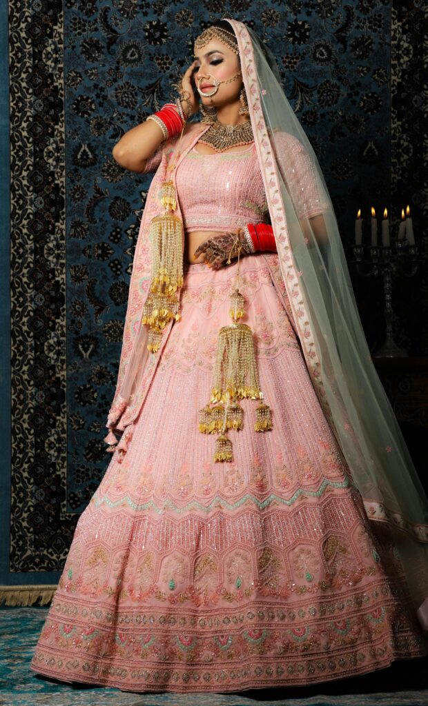 A radiant bride in a baby pink designer bridal lehenga with detailed embroidery, traditional jewellery, and kalire, posing gracefully — perfect for lehenga for women inspiration.