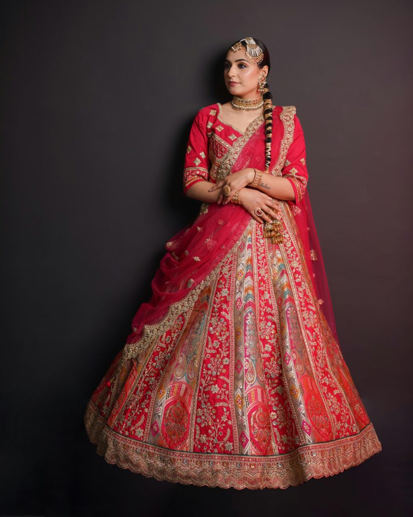 Bride in a cherry red traditional lehenga with multicolour woven patterns and gold embroidery, styled with a braid and jewellery — a stunning bridal lehenga for women.