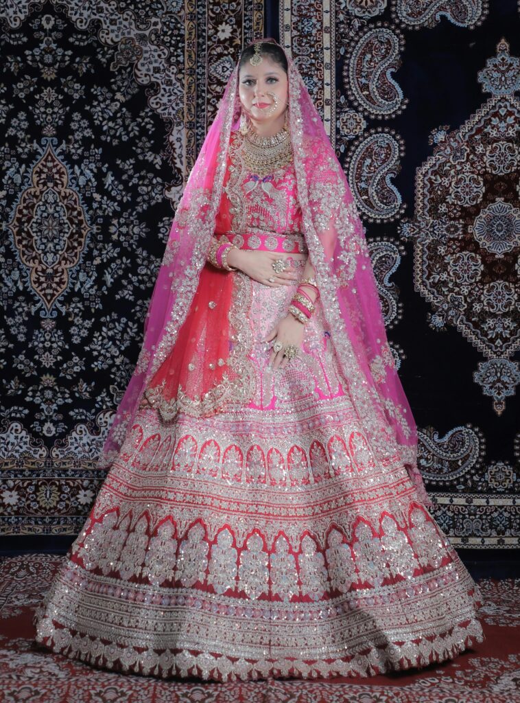 Bride wearing a stunning pink lehenga with intricate silver embroidery and dual-tone dupatta, posing against a royal carpet backdrop — ideal for showcasing bridal lehenga for women.