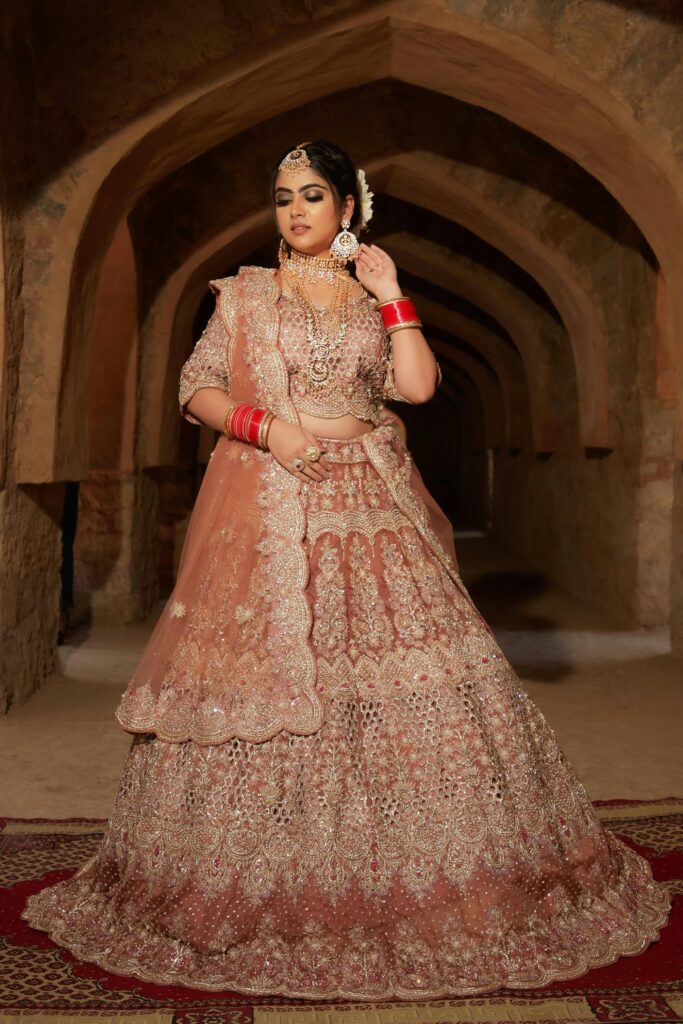 Bride wearing a peach-gold bridal lehenga with intricate embroidery and traditional red bangles, standing in an arched stone hallway — a stunning designer lehenga for women.